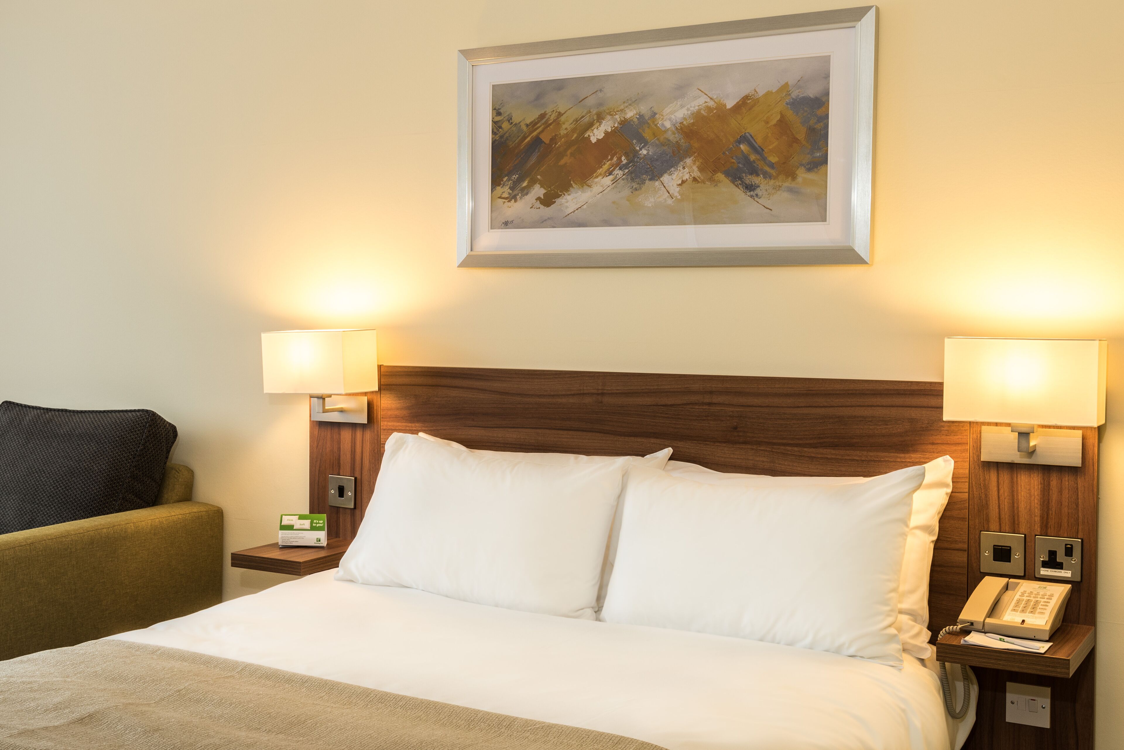 A hotel room with a neatly made bed, two bedside lamps, a telephone, and a framed abstract painting on the wall.
