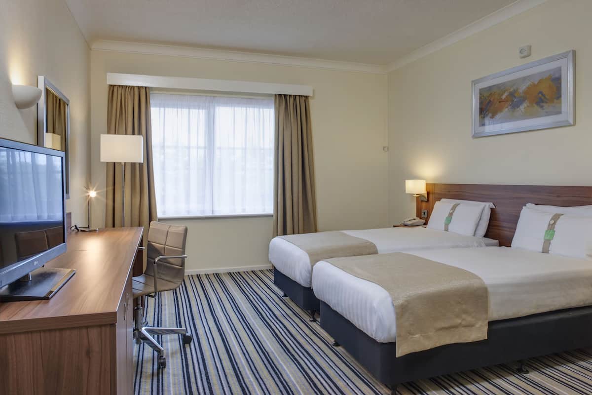 A hotel room with two beds, a desk with a computer, a chair, a window with curtains, and a painting on the wall.