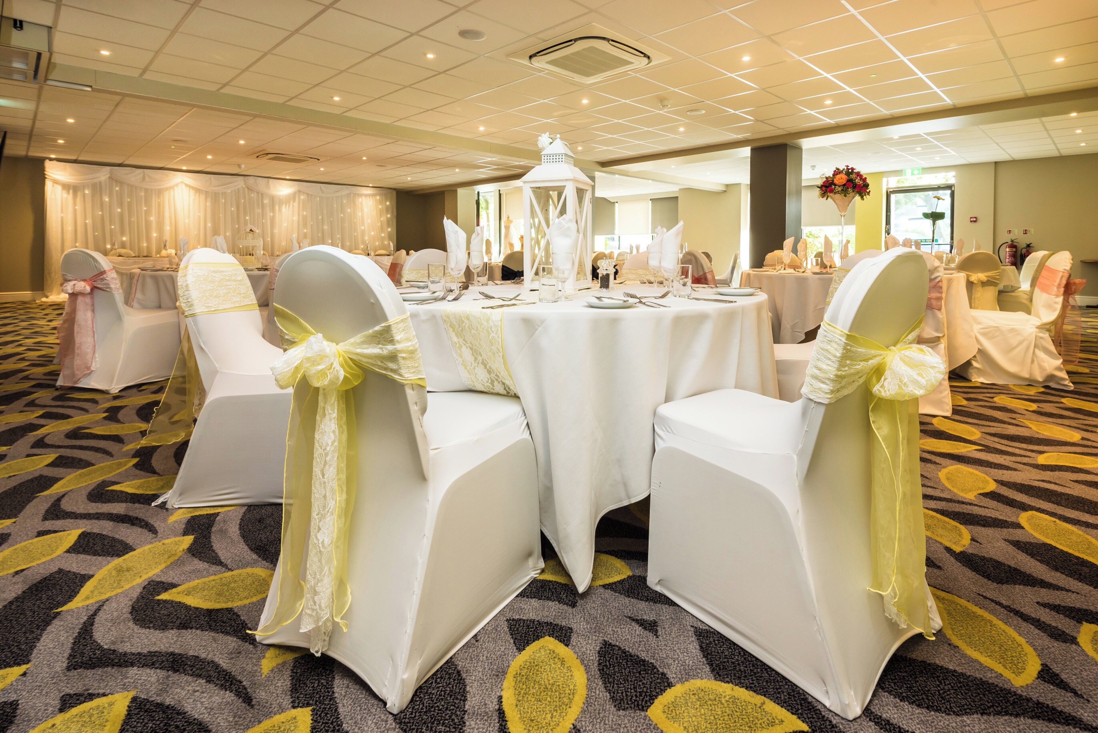 Un salón de banquetes con mesas redondas, sillas blancas con cintas amarillas y una alfombra estampada.