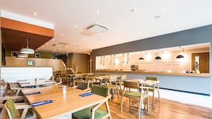 A modern restaurant with wooden tables and green cushioned chairs, a bar area, and a visible menu board. - Holiday Inn Lancaster by IHG (Lancaster)