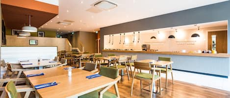 Un ristorante moderno con tavoli in legno e sedie imbottite verdi, un'area bar e una lavagna con il menu ben visibile.