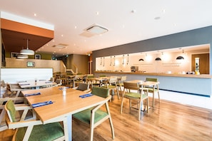 A modern restaurant with wooden tables and green cushioned chairs, a bar area, and a visible menu board. - Holiday Inn Lancaster by IHG (Lancaster)
