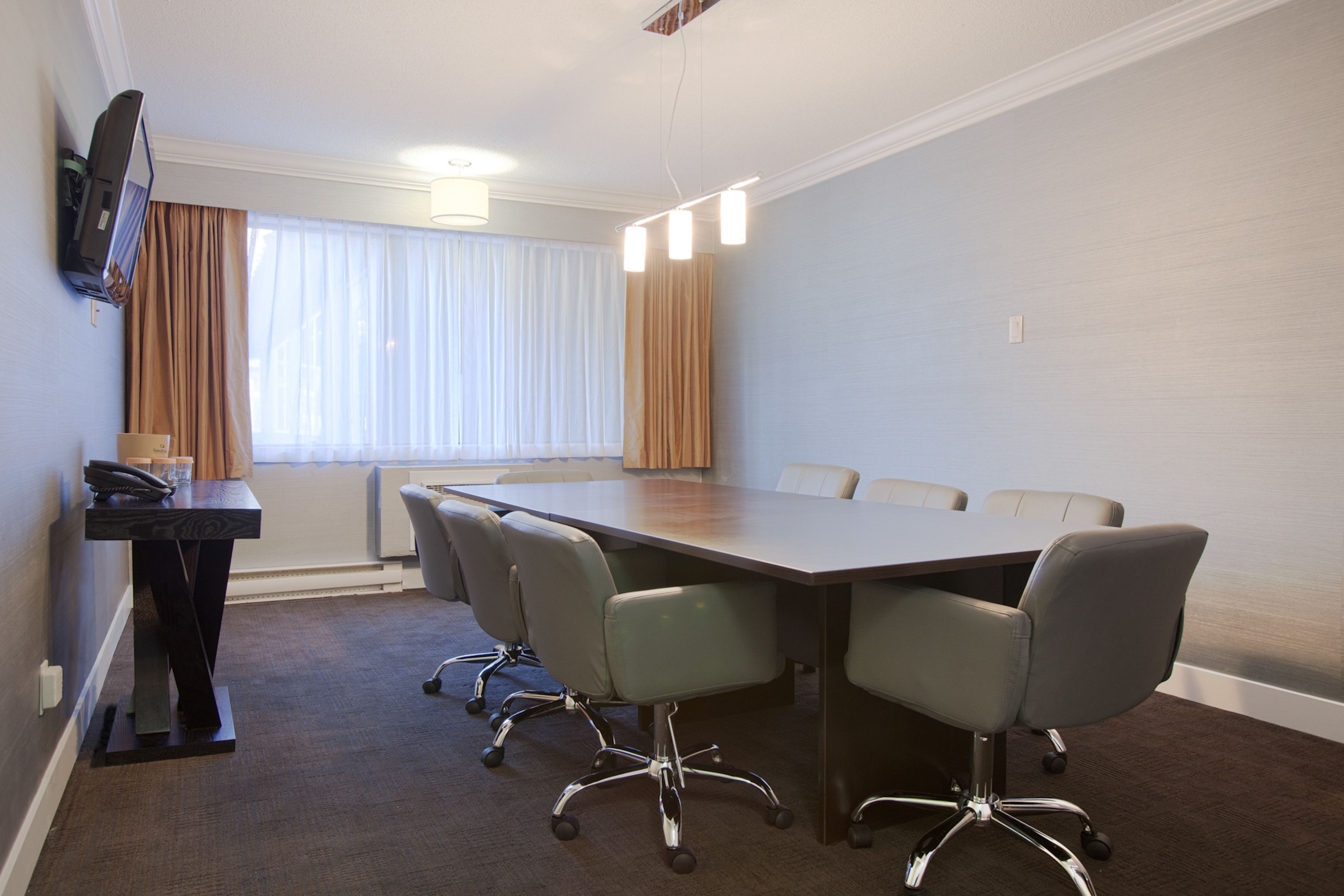 A conference room with a long table, four chairs, a small desk with a phone, and a TV mounted on the wall.