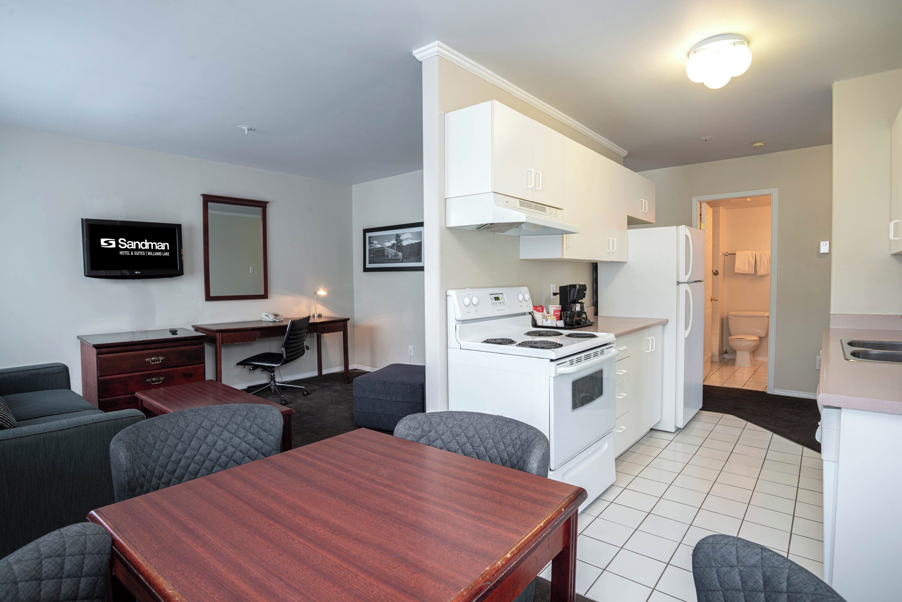 A compact kitchen with white appliances, a dining area with a wooden table, and a TV mounted on the wall.