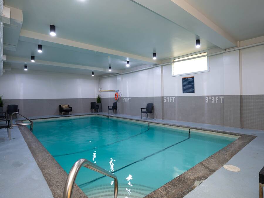 An indoor swimming pool with clear water, surrounded by a tiled floor and walls, with ceiling lights and a window on the right side.