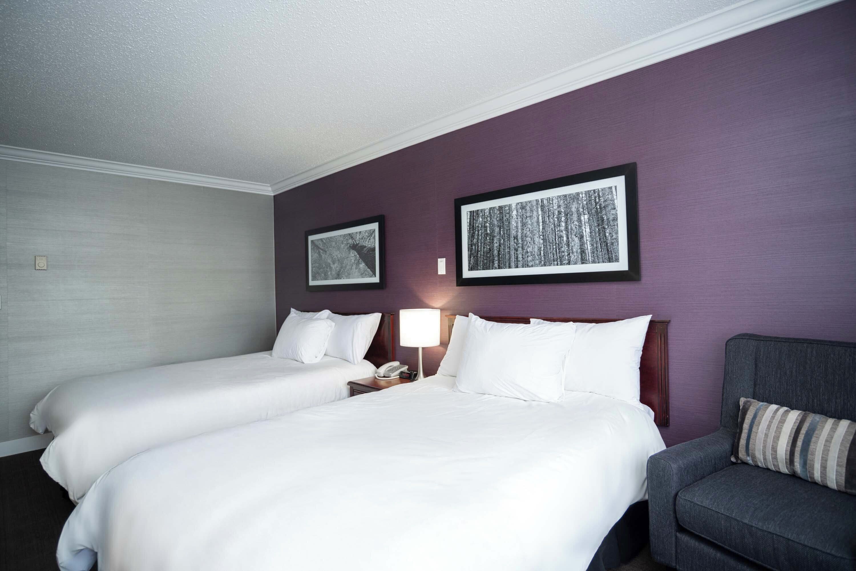 A hotel room with two beds, a nightstand with a lamp, and a chair.