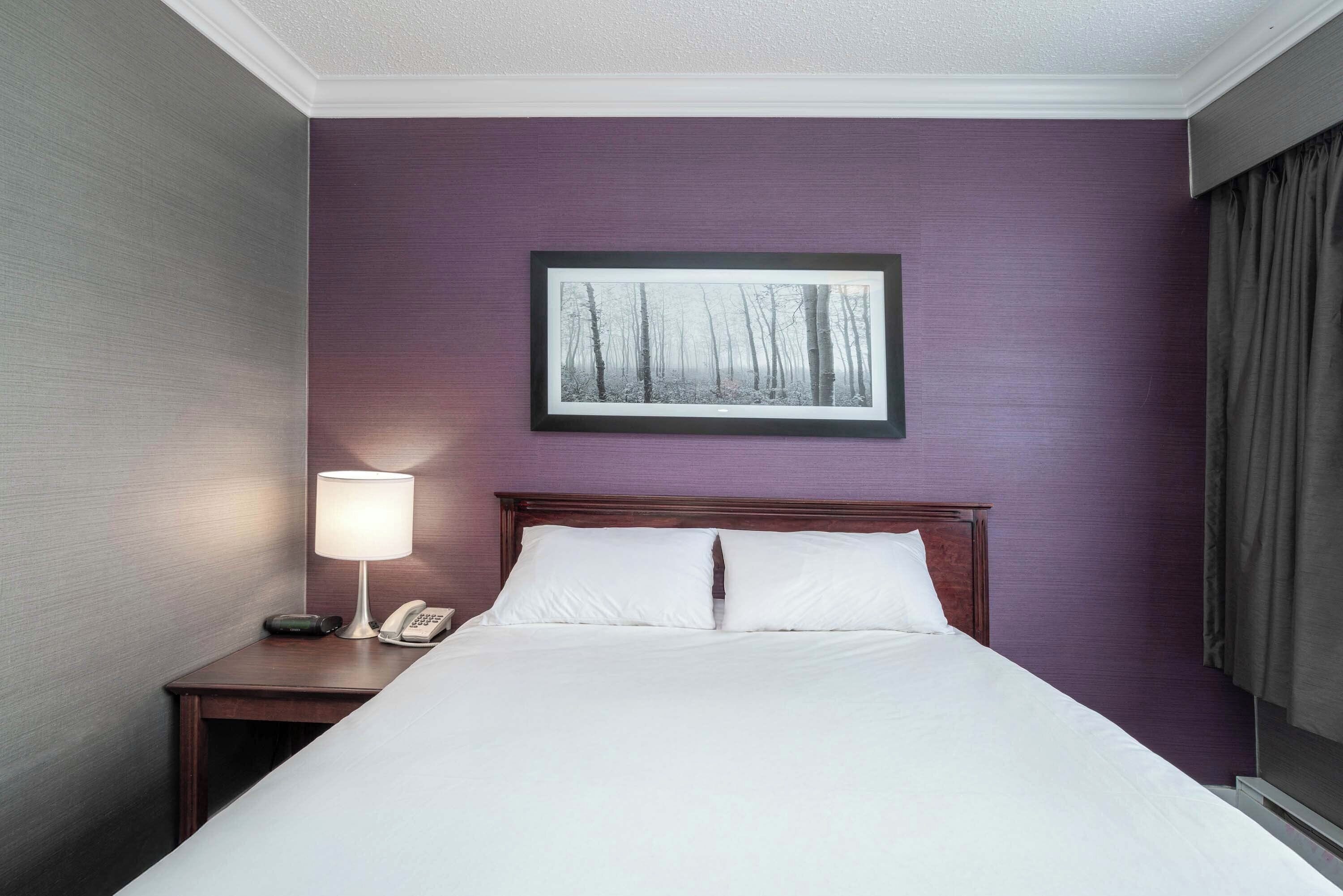 Una habitación de hotel con cama, mesita de noche con lámpara, teléfono y un cuadro en la pared.