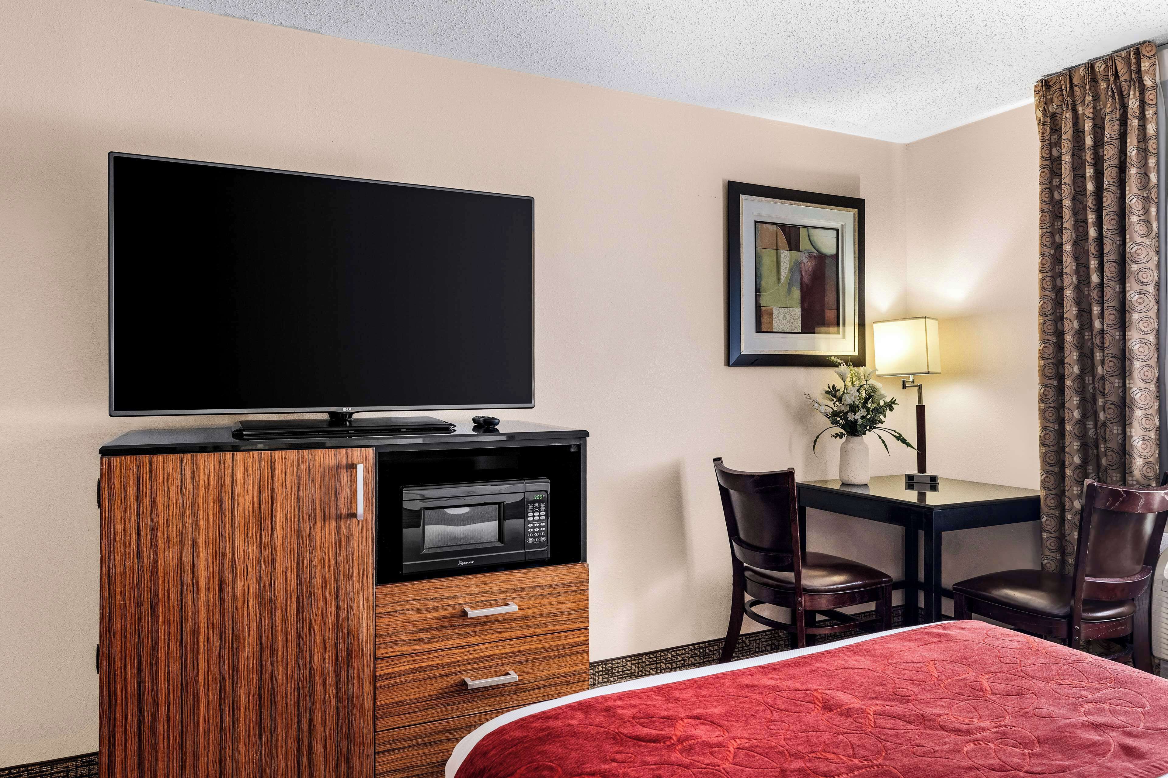 A hotel room with a flat-screen TV, a microwave, a desk with two chairs, a lamp, and a bed with a red bedspread.