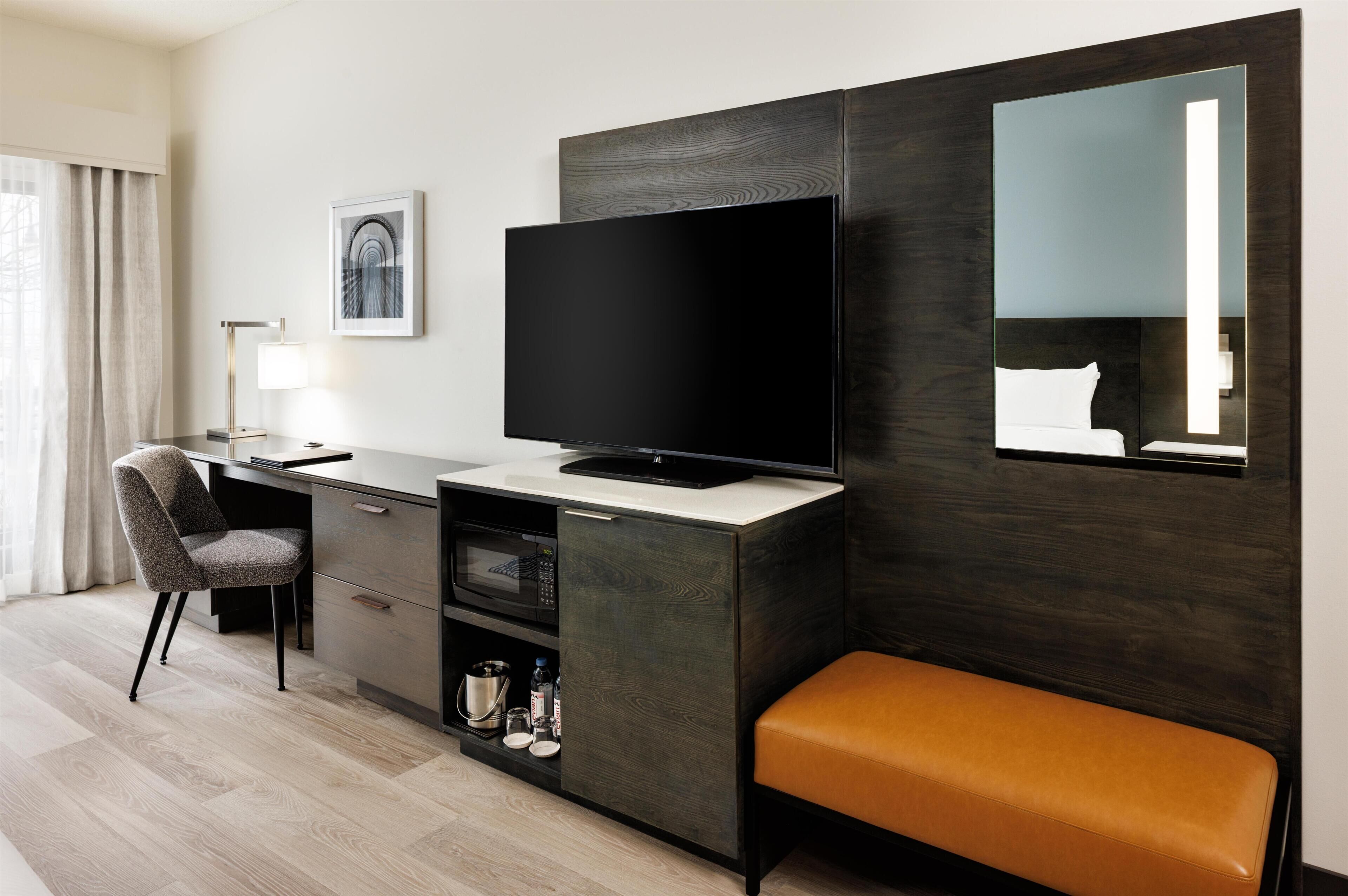 Una habitación de hotel moderna con una televisión de pantalla plana, un escritorio con una silla, una pequeña cocina y un banco, además de un espejo grande.