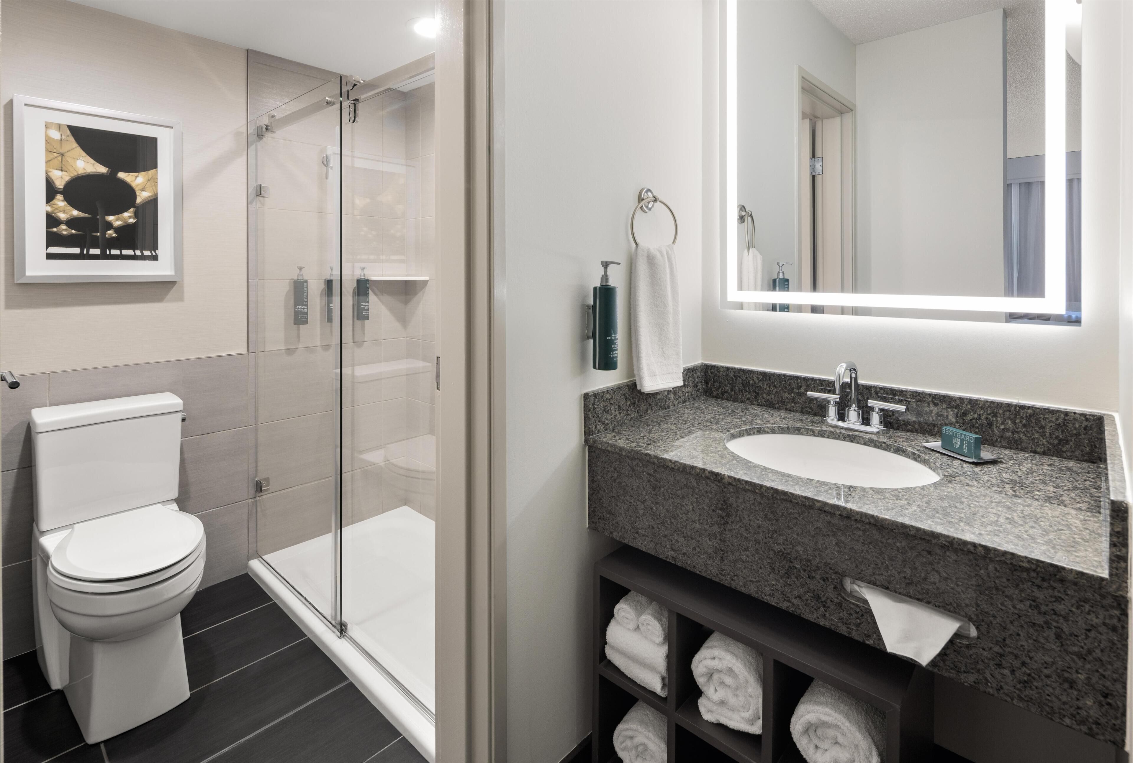 Un baño moderno con ducha envuelta en vidrio, inodoro blanco, lavabo de granito y un espejo grande.