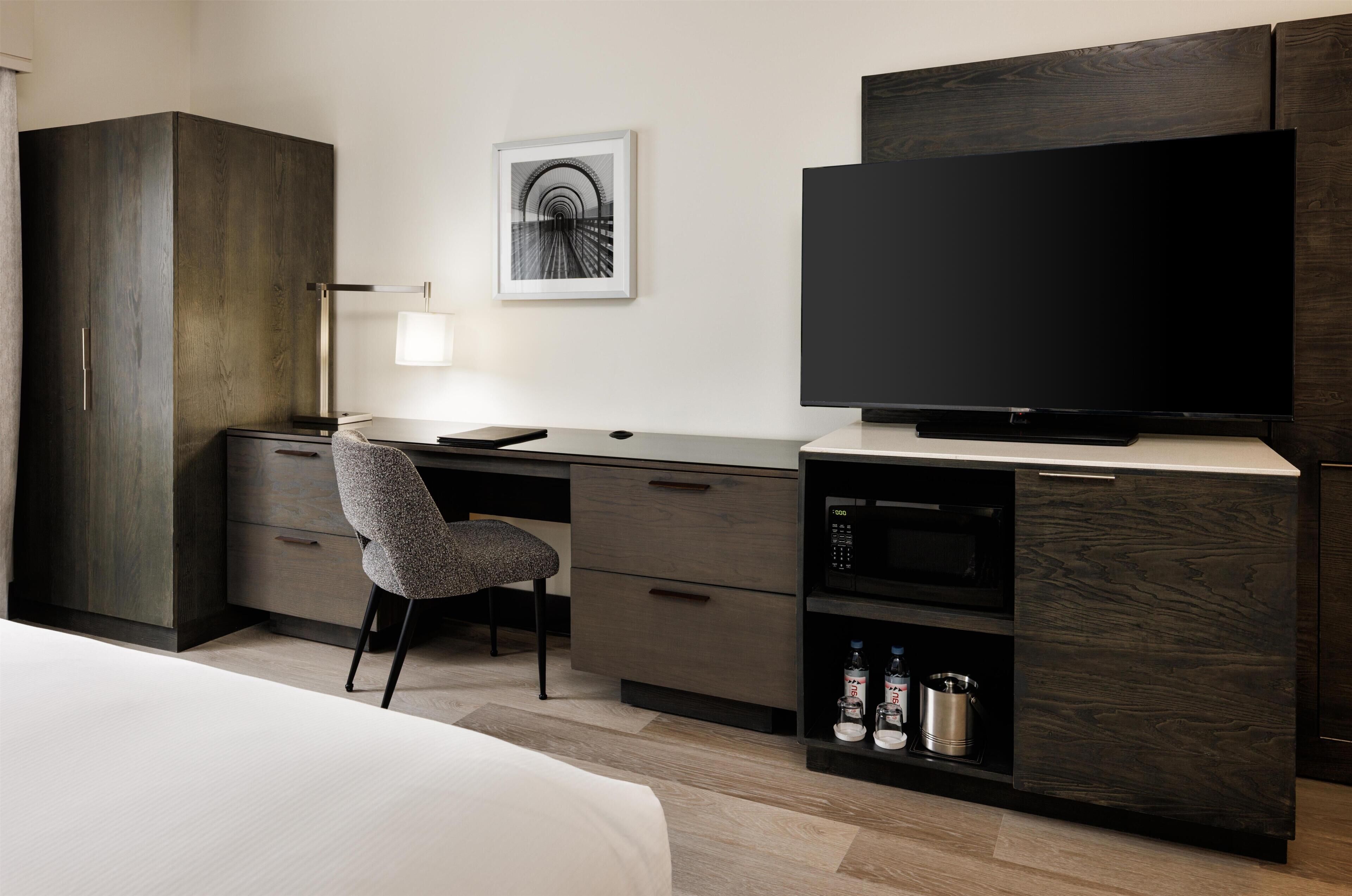 Et moderne hotelværelse med en stor seng, et skrivebord med stol, et fladskærms-TV, en lampe og et billede i ramme på væggen.