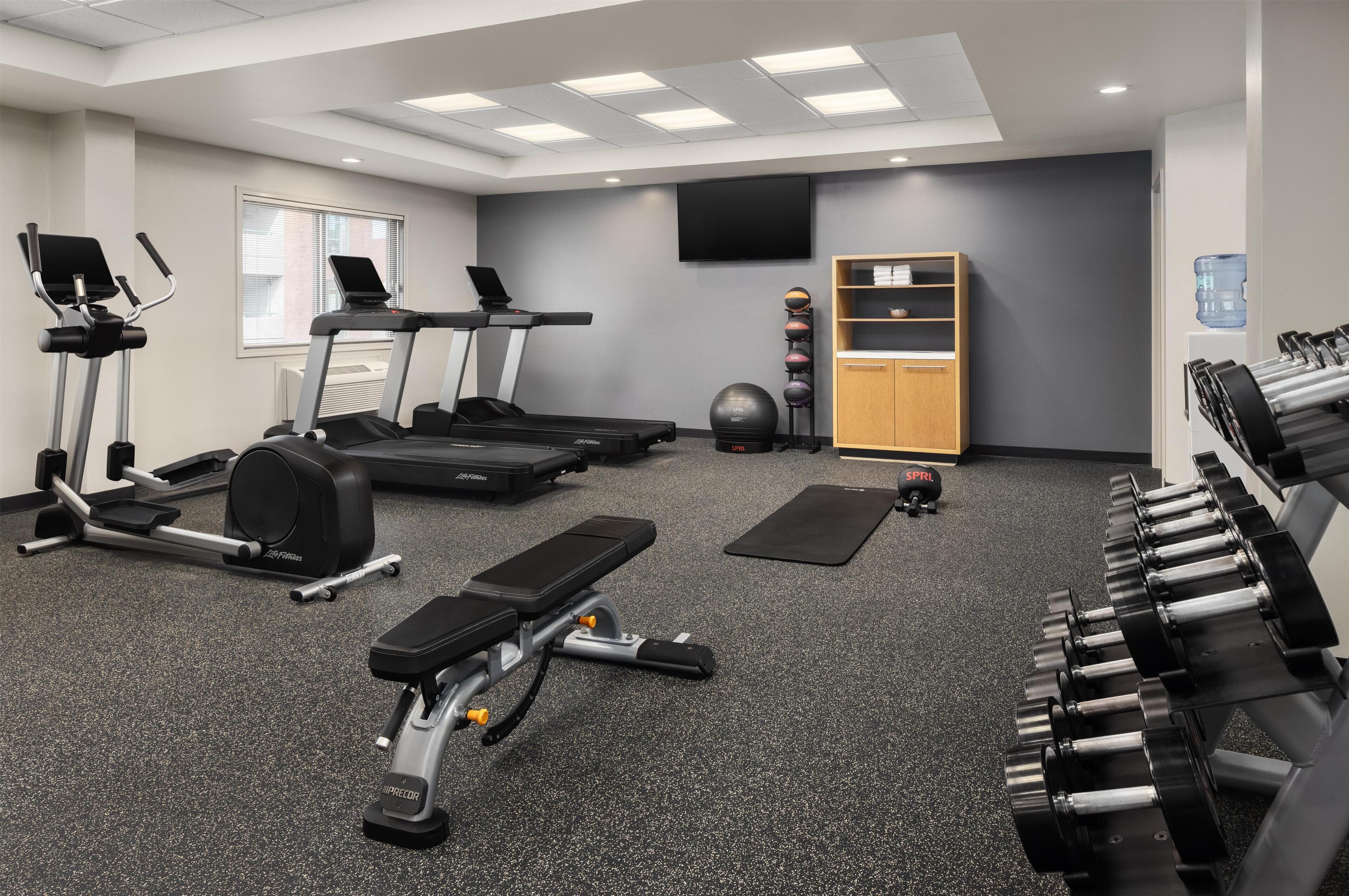 A gym with treadmills, elliptical machines, a bench press, dumbbells, a water bottle, and a television on the wall.