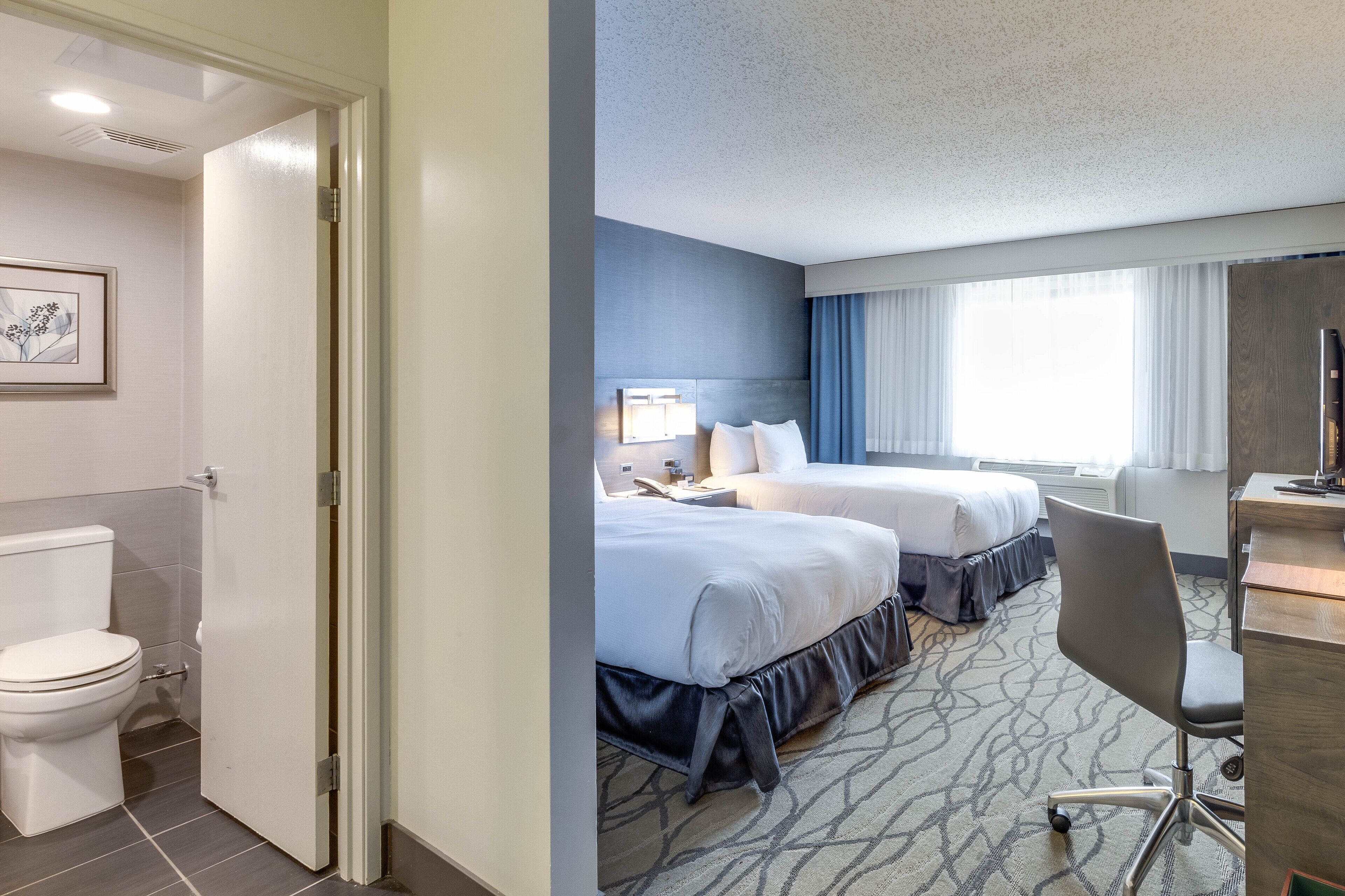 A hotel room with two beds, a desk, a chair, a toilet, and a framed picture on the wall.