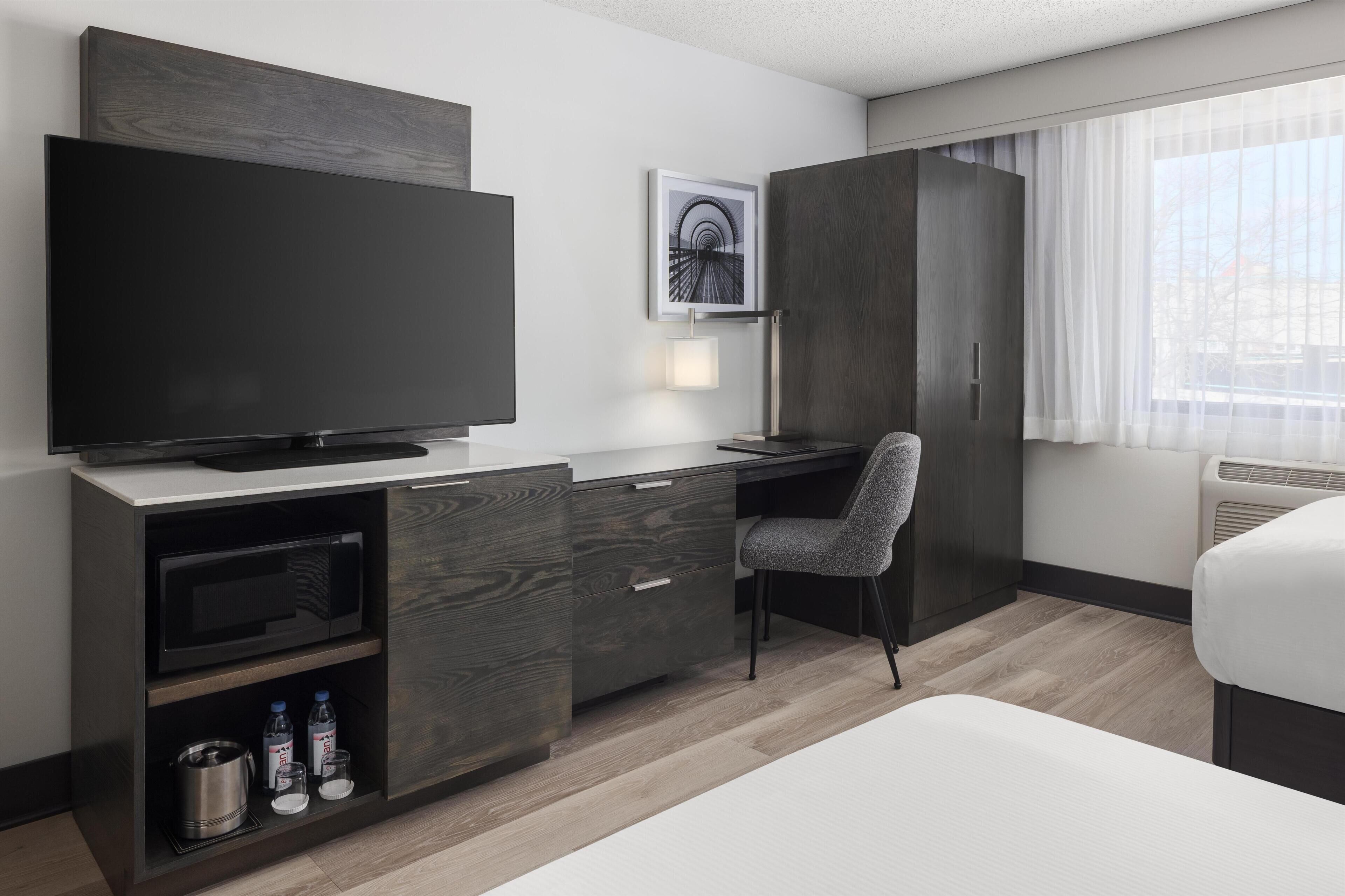 A modern hotel room with a flat-screen TV, a desk, a chair, a bed, a wardrobe, and a window with curtains.