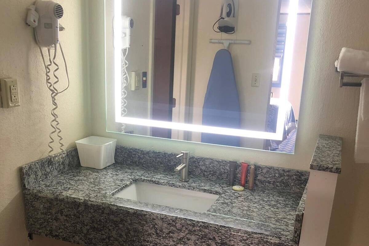 A bathroom with a granite countertop, a sink, a mirror with a light, a hairdryer, and a trash bin.