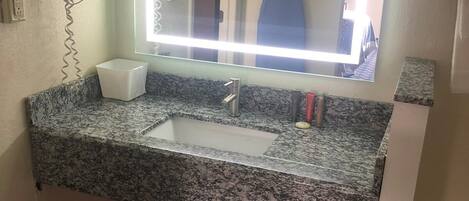 A bathroom with a granite countertop, a sink, a mirror with a light, a hairdryer, and a trash bin.