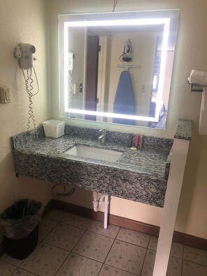 A bathroom with a granite countertop, a sink, a mirror with a light, a hairdryer, and a trash bin. - Aderi Hotel Lewisburg (Lewisburg)
