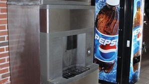 Un dispensador de bebidas de acero inoxidable con el logotipo de Pepsi, situado en un pasillo con pared de ladrillo y suelo de baldosas.