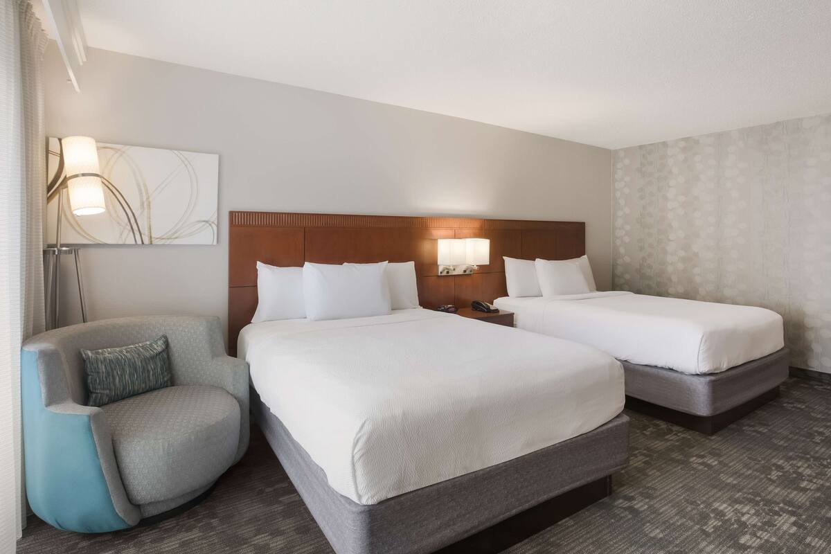A hotel room with two beds, a chair, a nightstand, and a wall-mounted lamp.