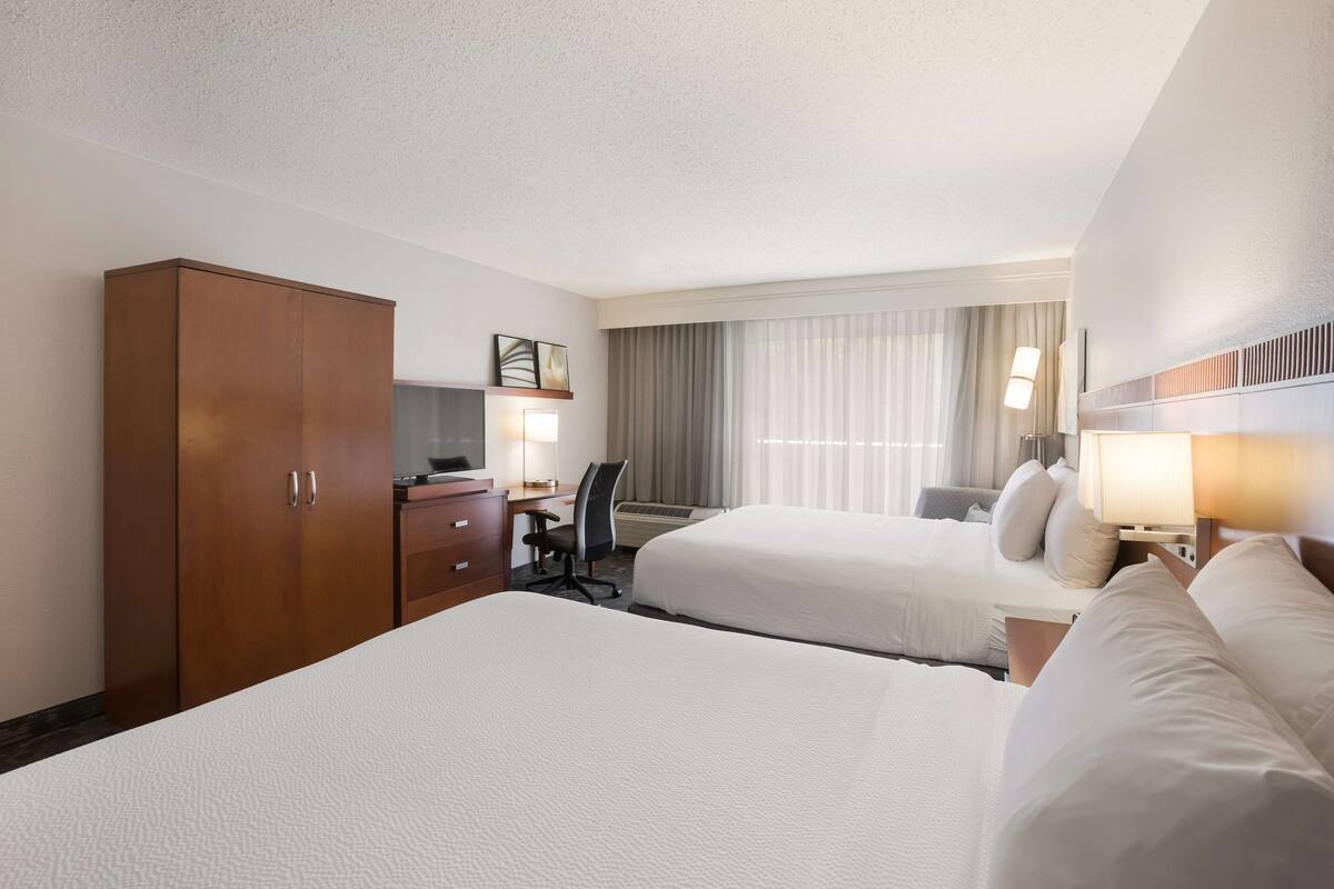 A hotel room with two beds, a desk with a chair, a wardrobe, and a window with curtains.