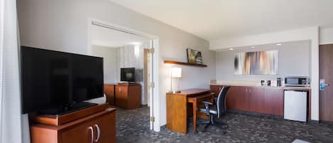 Habitación de hotel con televisor, escritorio de madera, silla de oficina, kitchenette y una cama.