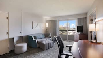 Une chambre d’hôtel équipée d’un canapé, d’un pouf, d’un fauteuil, d’un bureau, d’une télévision et d’une grande fenêtre donnant sur la ville.