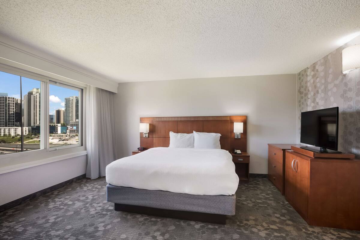 A hotel room with a large bed, a TV, a wooden nightstand, and a view of the city.