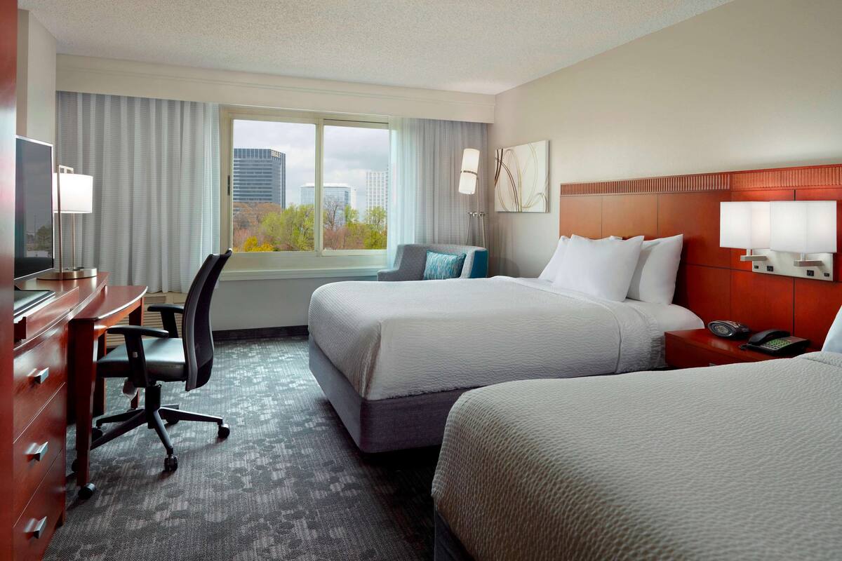 A hotel room with two beds, a desk with a chair, a TV, and a window with a city view.
