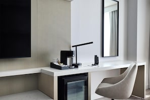 A modern hotel room with a white desk, a black coffee maker, a lamp, and a grey chair.
