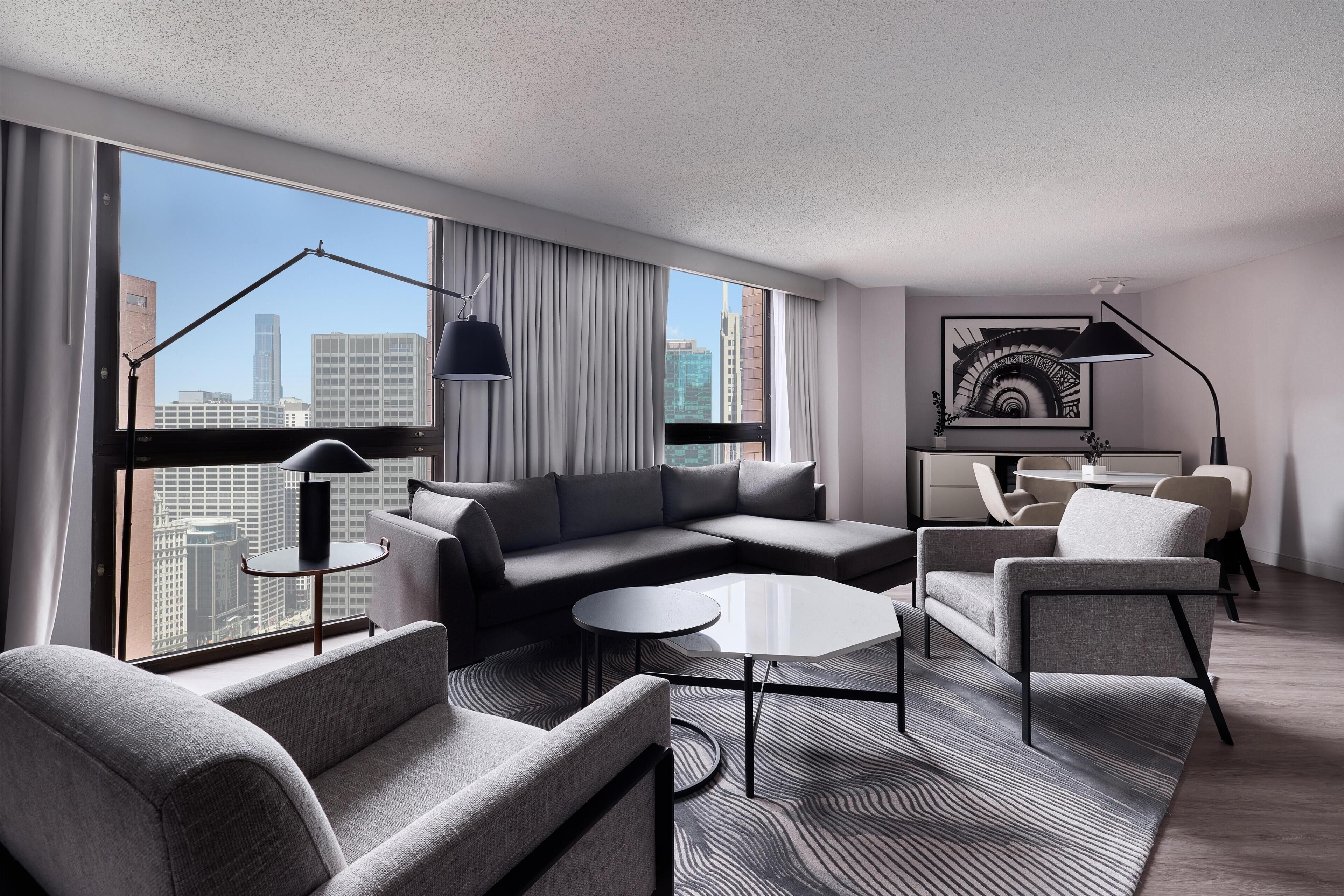 A modern hotel room with a sectional sofa, a round coffee table, and a dining area with chairs.