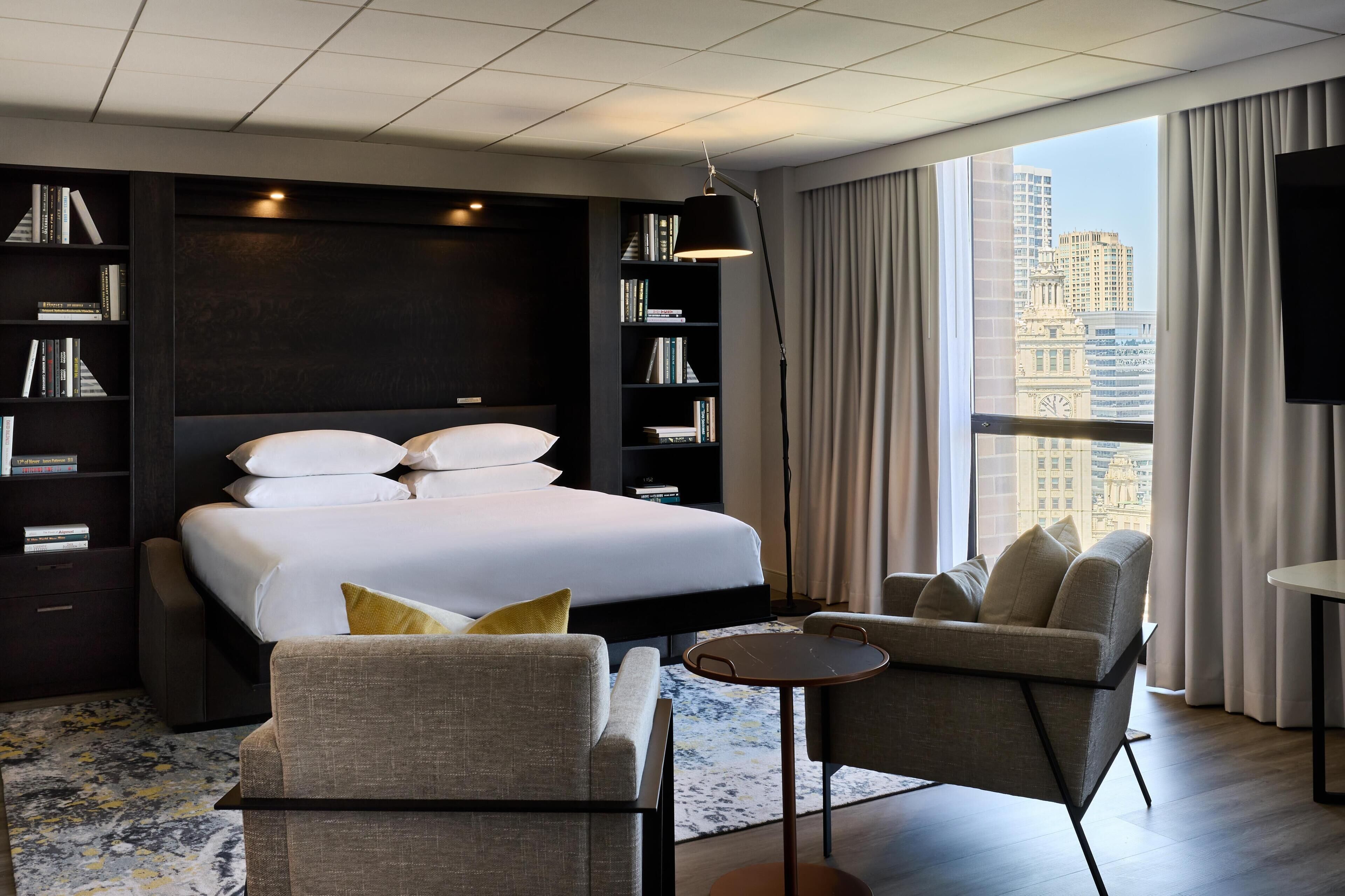 A modern hotel room with a large bed, two armchairs, a small table, and a bookshelf.