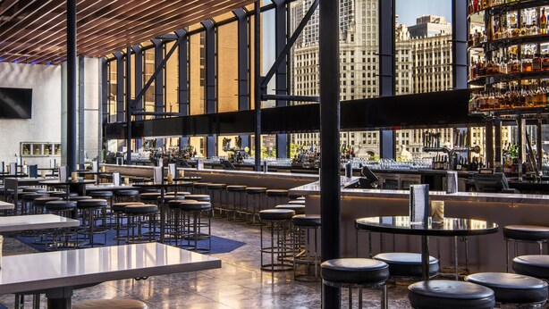 A spacious bar area with high ceilings and large windows, offering views of a cityscape.