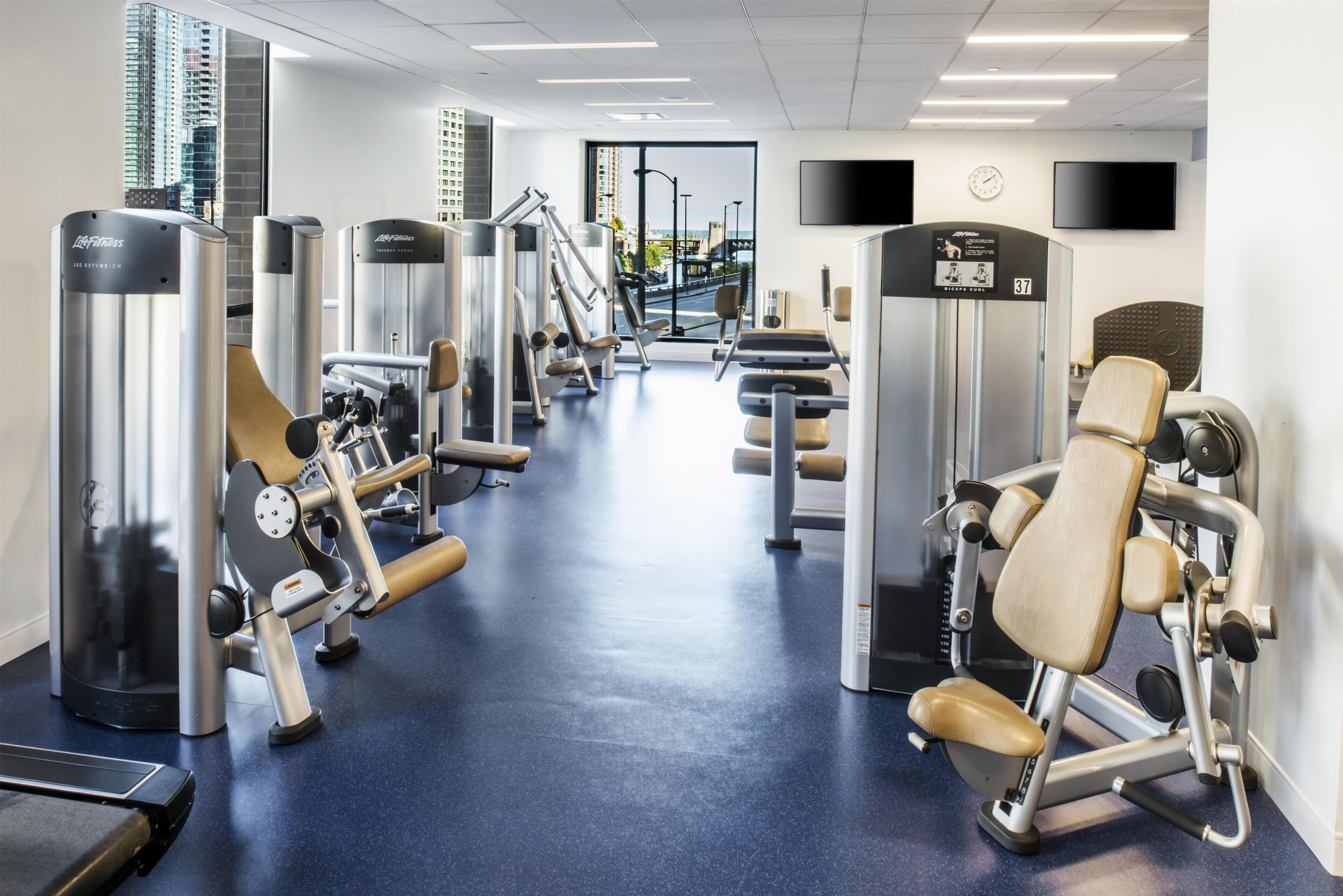 Ett gym med olika träningsmaskiner, ett stort fönster med utsikt över staden och en klocka på väggen.