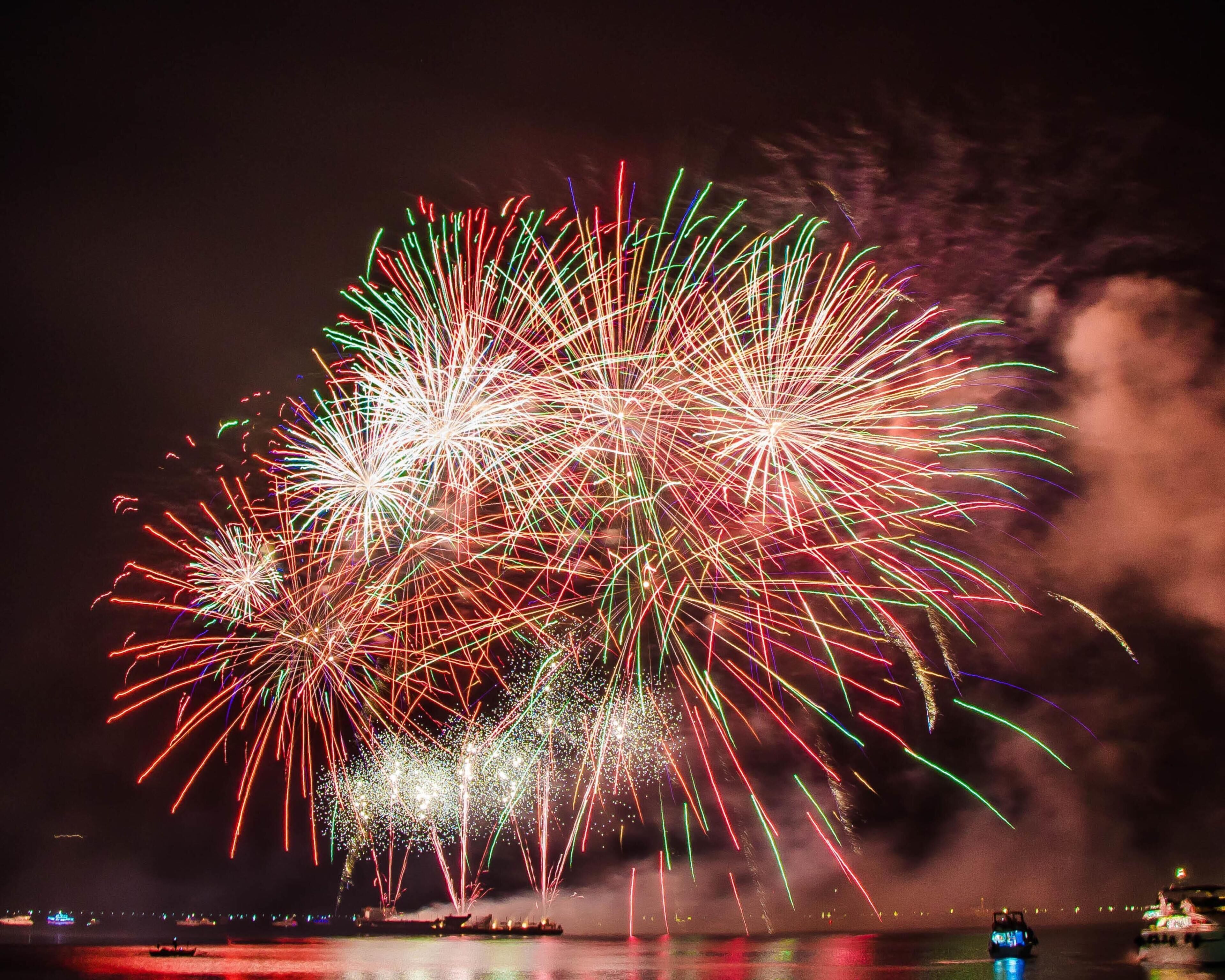 A nighttime view of fireworks over a body of water with boats and distant city lights.