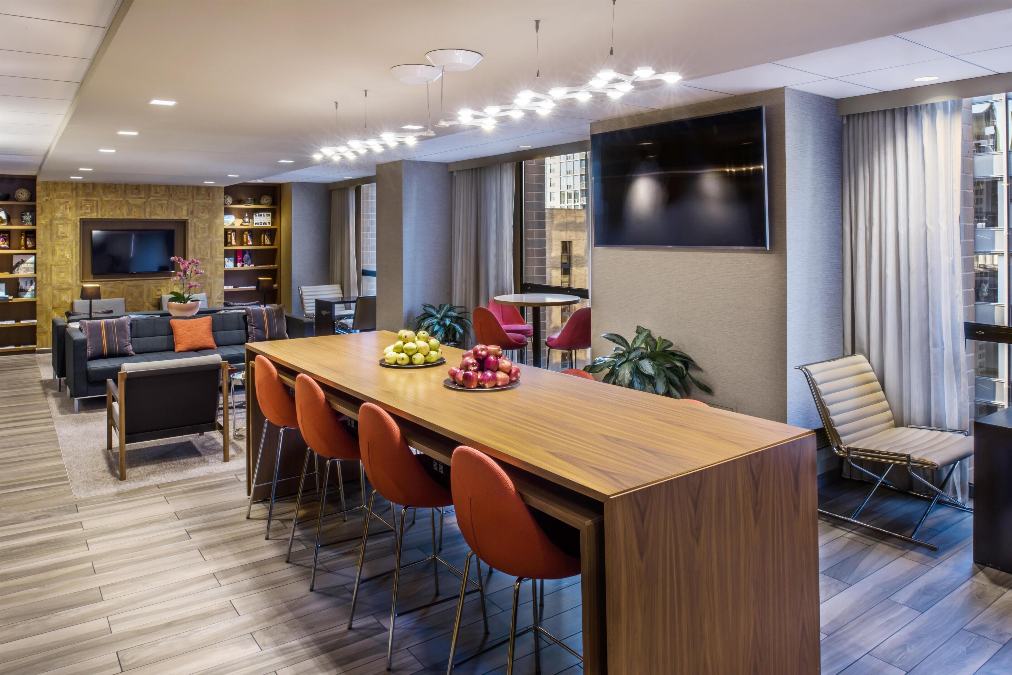 A modern hotel lobby with a wooden reception desk, red chairs, a sofa, and a TV mounted on the wall.