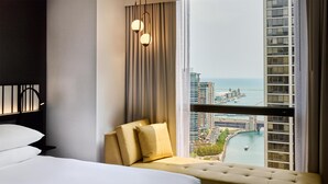 A hotel room with a bed, a sofa with cushions, a large window with a view of a cityscape and water.