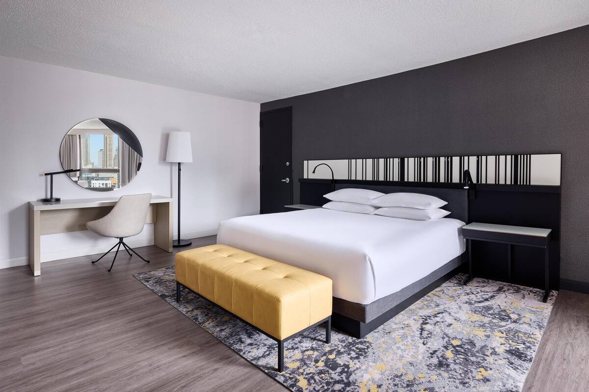 A modern hotel room with a bed, a desk, a chair, a round mirror, and a bench.