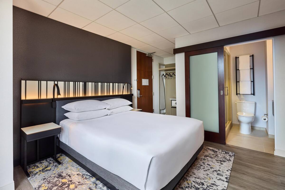 A hotel room with a large bed, bedside table, a mirror, a wardrobe, and a bathroom with a toilet and shower.
