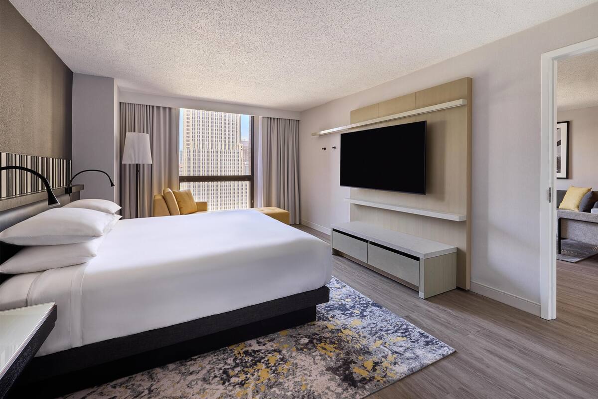 A modern hotel room with a large bed, a flat-screen TV, a desk, and a window with a city view.