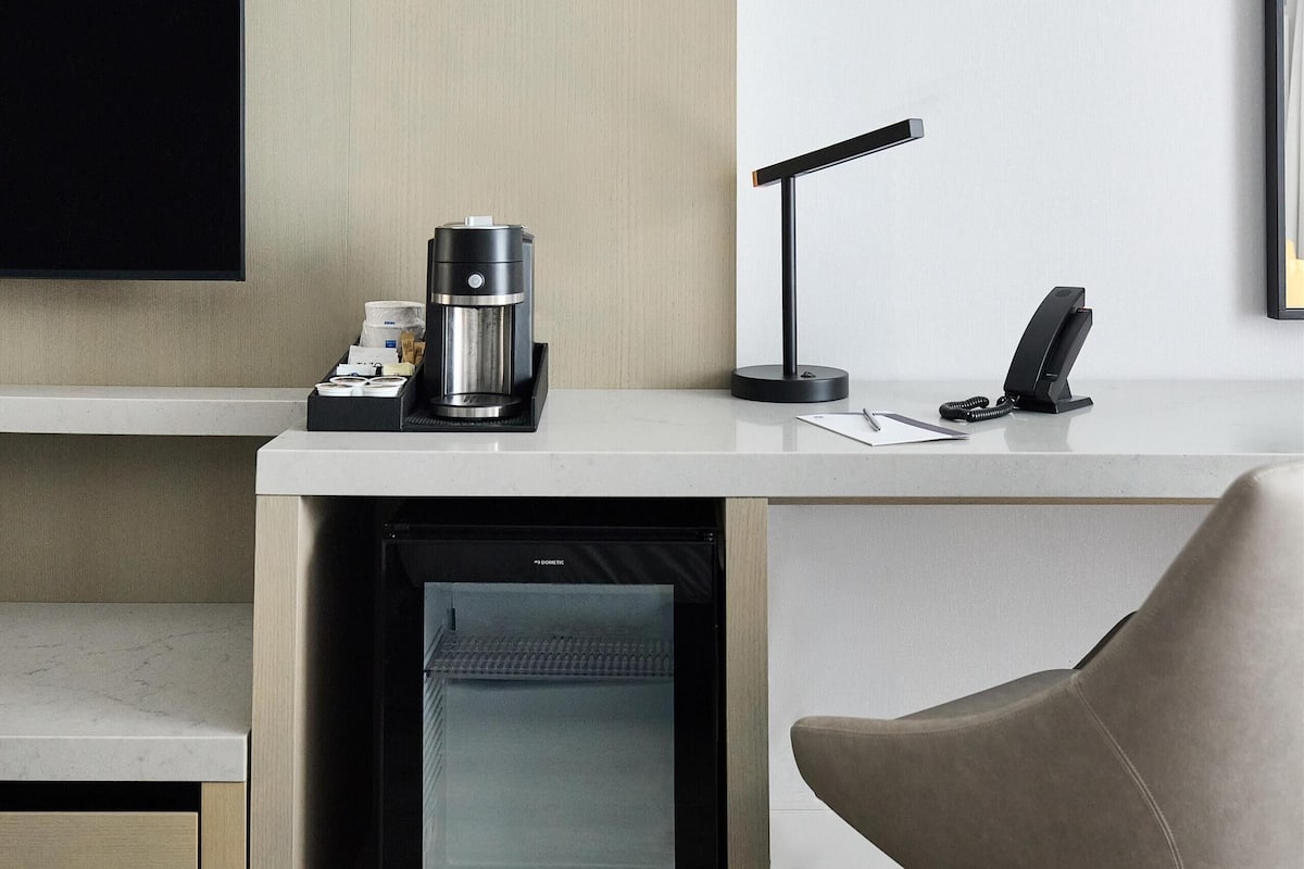 A modern hotel room with a flat-screen TV, a coffee machine, a desk lamp, a phone, and a mini-fridge.