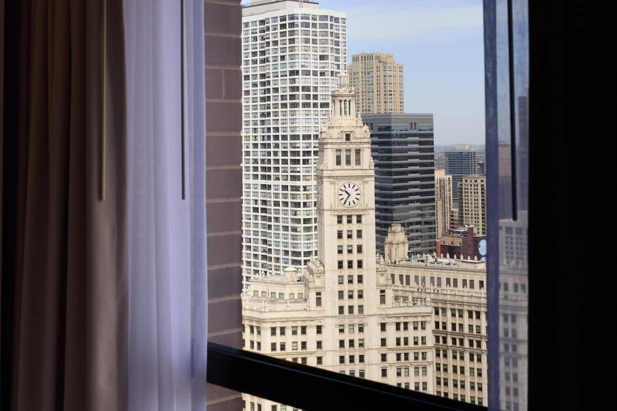 A view of a cityscape through a window with curtains, featuring a prominent clock tower and modern high-rise buildings.