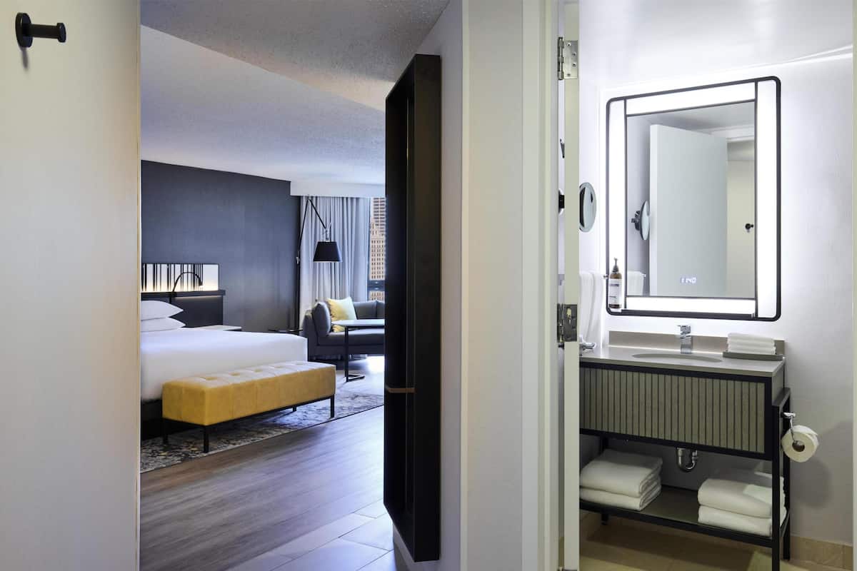 A hotel room with a bed, a bench, a sofa, a large mirror, and a bathroom with a sink and towel rack.