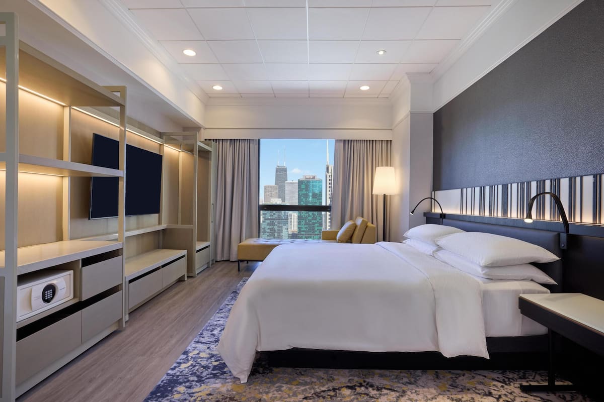 A hotel room with a bed, bedside table, lamp, TV, and a view of the city skyline.