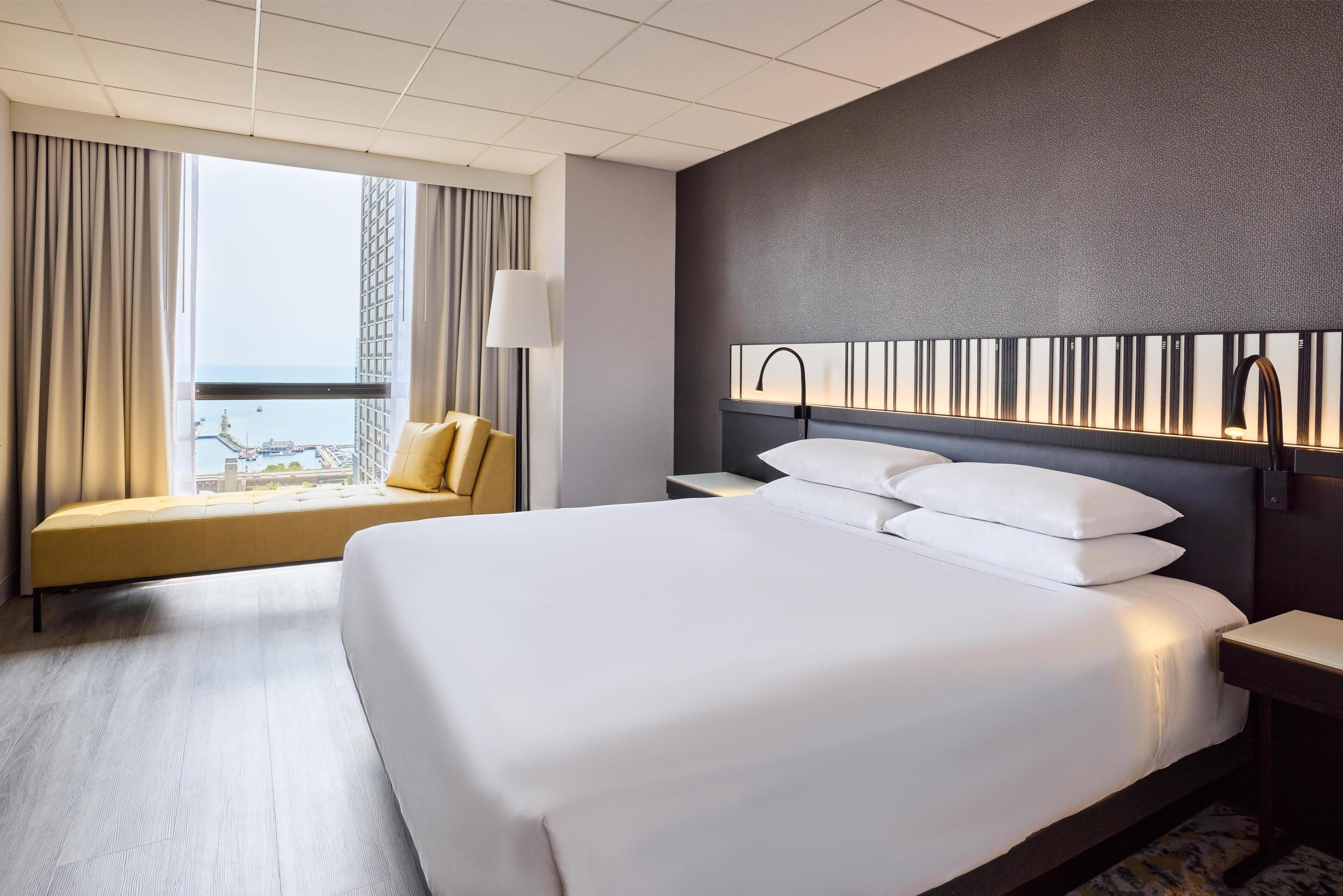 A hotel room with a large bed, a sofa, a nightstand, a lamp, and a view of the city.