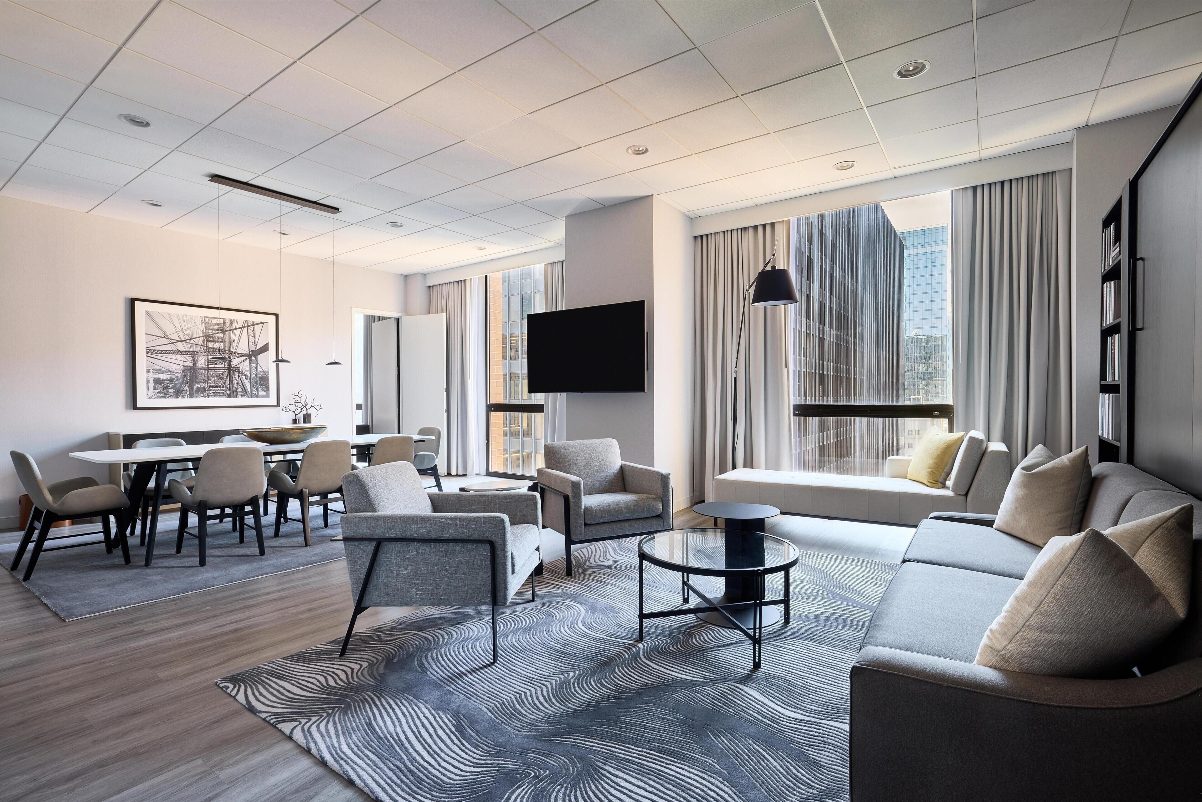 Una habitación de hotel moderna con comedor, un conjunto de sofás, un televisor de pantalla plana y ventanas grandes con cortinas.
