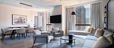 Una habitación de hotel moderna con comedor, un conjunto de sofás, un televisor de pantalla plana y ventanas grandes con cortinas.