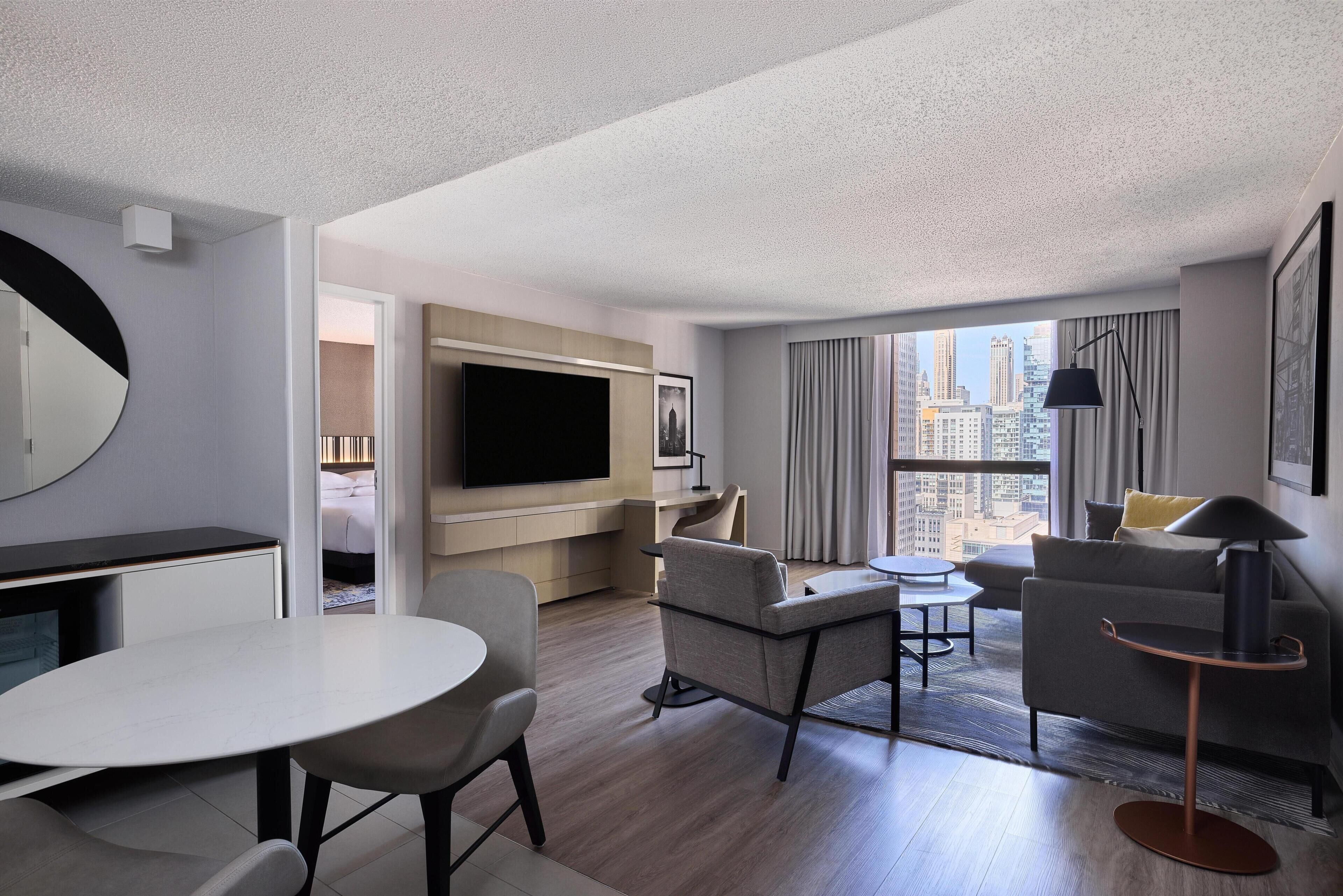 A modern hotel room with a flat-screen TV, a dining area, a sofa, and a large window offering a city view.
