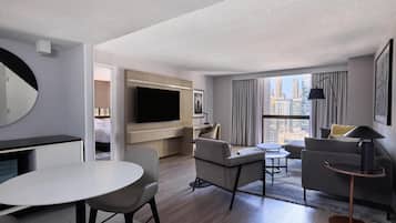 A modern hotel room with a flat-screen TV, a dining area, a sofa, and a large window offering a city view.