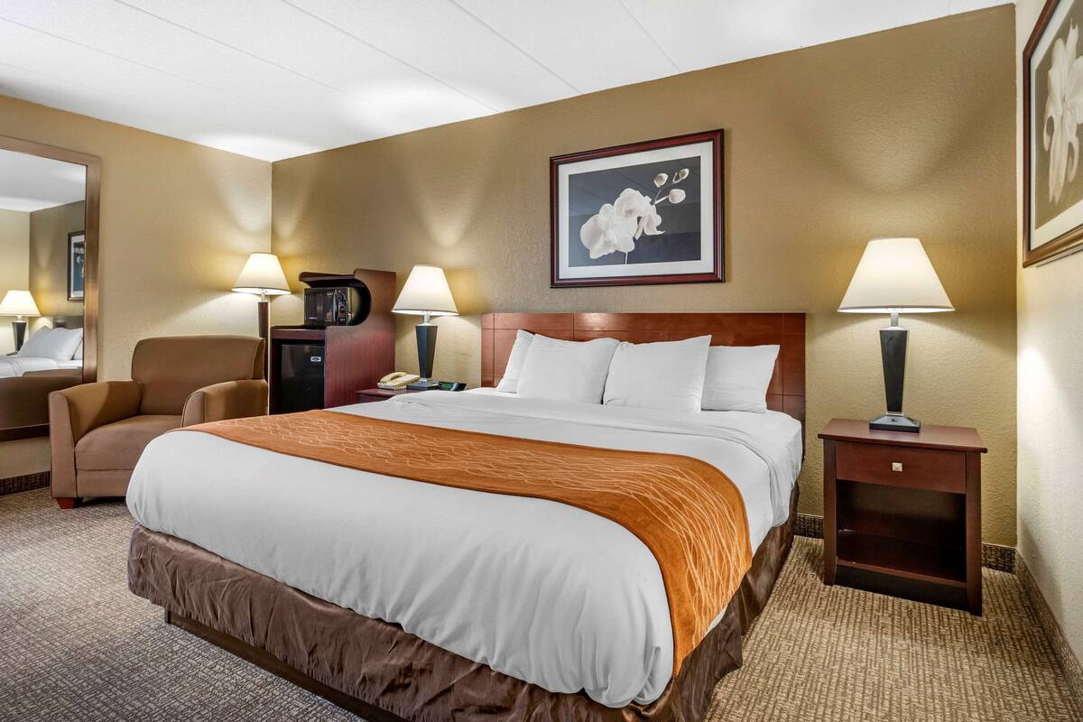 A hotel room with a large bed, two bedside lamps, a chair, a desk with a telephone, and a framed picture on the wall.