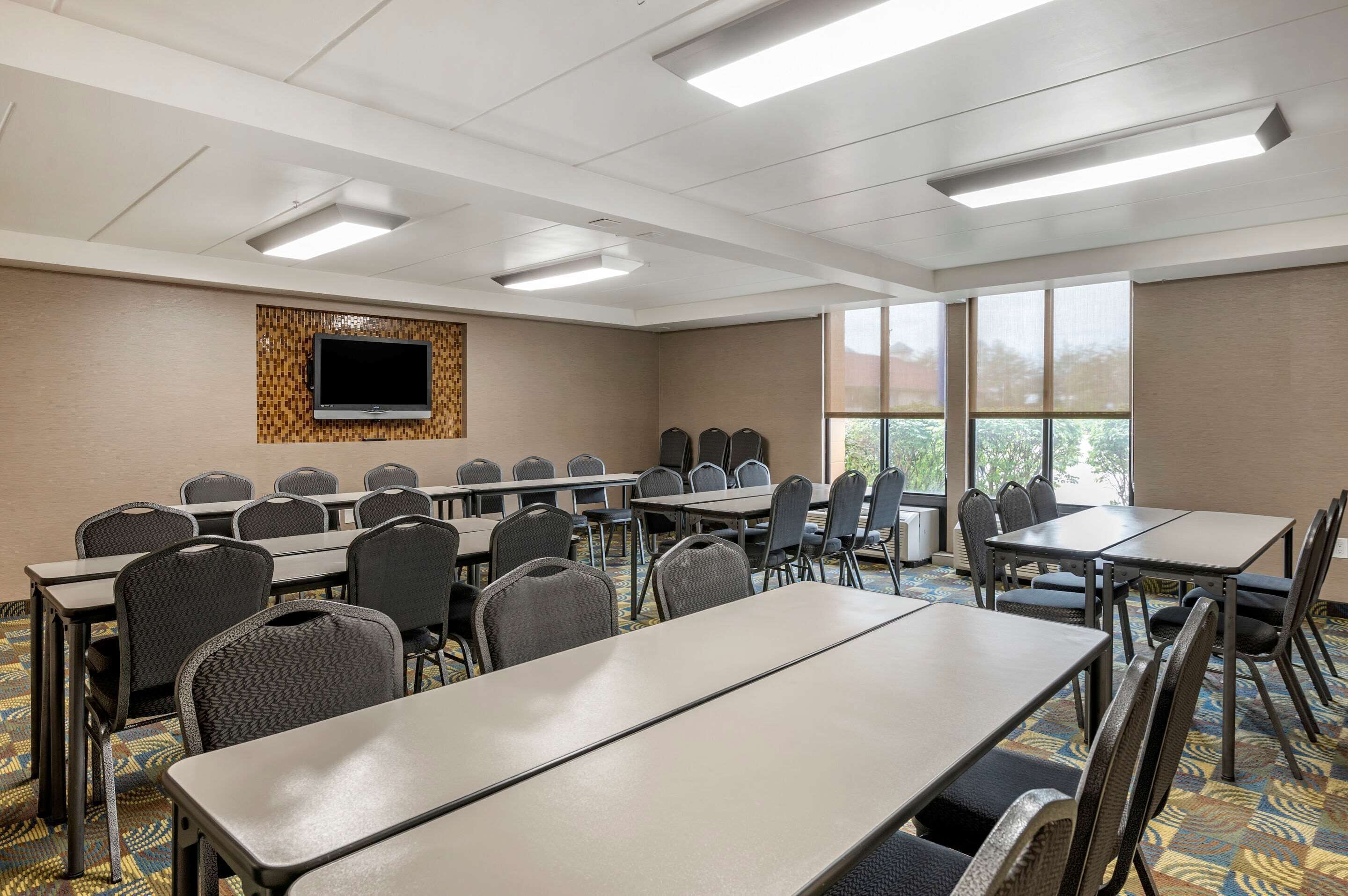 A conference room with multiple tables and chairs, a flat-screen TV mounted on the wall, and large windows providing natural light.